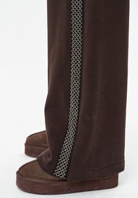 Bruine joggingbroek met een gestructureerde zilveren accenten langs de zijkant, gecombineerd met bruine slippers met een suède-achtige textuur.