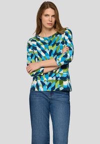 Frau mit hellbraunen Haaren, die ein langärmliges Oberteil mit blau-grün-weißem geometrischem Muster und blaue Jeans trägt, Arme verschränkt, neutraler Hintergrund.