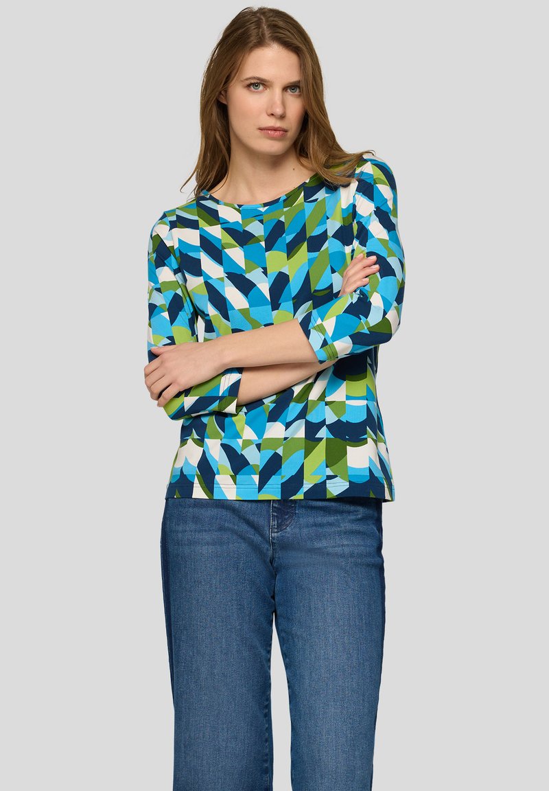 Frau mit hellbraunen Haaren, die ein langärmliges Oberteil mit blau-grün-weißem geometrischem Muster und blaue Jeans trägt, Arme verschränkt, neutraler Hintergrund.