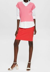 Vaaleanpunainen ribatoitu neuleliivi valkoisen nappipaidan päällä, yhdistettynä punaiseen kynähameeseen. Valkoiset nauhakorkokengät täydentävät asukokonaisuutta.