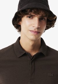 Brun polo-shirt med ett texturerat tyg och en klassisk krage. Har en liten broderad logotyp på bröstet och en svart kordflöjls bucket hat.