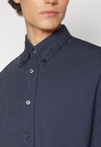 Hombre joven con una camisa de pana azul oscuro, abotonada, con el cuello abotonado, visible desde el cuello hasta el pecho.