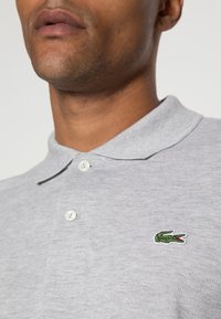 Graues Poloshirt aus strukturiertem Stoff, mit einem kleinen grünen gestickten Krokodil-Logo und zwei weißen Knöpfen am Kragen.