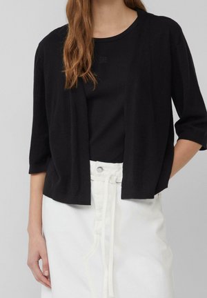 Femme portant un cardigan noir ouvert devant sur un top noir et un pantalon blanc taille haute avec une ceinture en tissu nouée devant.