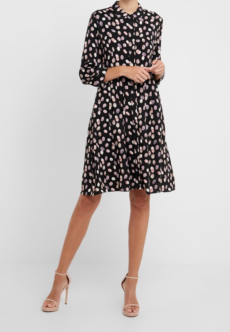 Femme portant une robe noire longueur genou avec des taches abstraites roses et blanches, manches longues, devant boutonné, et sandales à talons hauts couleur nude.
