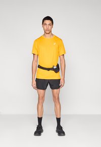 Πορτοκαλί T-shirt που απομακρύνει την υγρασία, συνδυασμένο με μαύρο σορτς για τρέξιμο, με μαύρη ζώνη μέσης που διαθέτει θήκη για μπουκάλι νερού, σχεδιασμός στενής εφαρμογής.