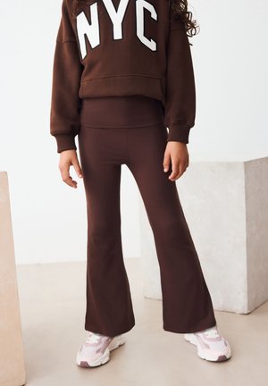 Brązowa bluza z dużymi białymi literami "NYC", zestawiona z brązowymi legginsami o rozszerzanych nogawkach i różowymi trampkami. Materiał o fakturze, casualowy design.