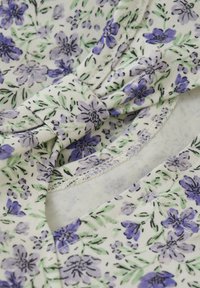 Blumengemusterter Stoff in Weiß mit lila Blumen und grünen Blättern. Ein geknotetes Detail am Halsauschnitt sorgt für Textur. Weiches Material.