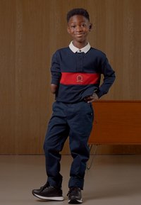 Granatowa koszulka polo z długim rękawem z czerwoną paską i logo, w połączeniu z granatowymi spodniami i czarnymi trampkami. Gładkie tekstury, swobodny krój i zorganizowany design.