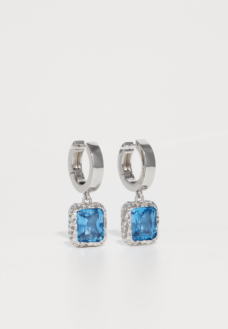 Silberne Creolen mit gehämmerten quadratischen Rahmen, die facettierte leuchtend blaue Edelsteine halten, gezeigt vor einem weißen Hintergrund.