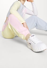 Bílé tenisky FILA s masivní podrážkou a černým logem, kombinované s pastelovými sportovními kalhotami v růžové a žluté barvě a světle modrým tričkem s dlouhým rukávem.