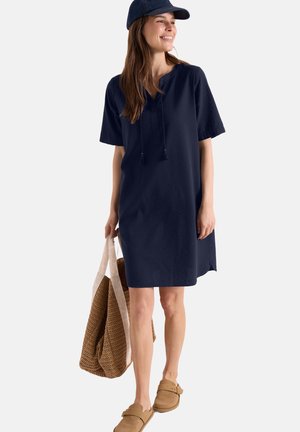 Vrouw glimlachend, draagt een marineblauwe jurk tot op de knie en een pet, draagt een grote geweven draagtas en draagt beige instappers.