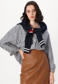 Camicia a righe con maniche larghe, abbinata a una gonna di pelle marrone. Un maglione di maglia blu è appoggiato attorno al collo con accenti rossi e bianchi.