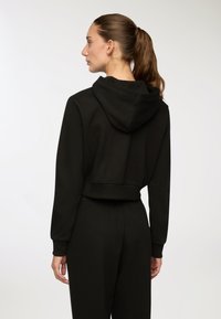 Donna con ponytail castano, che indossa un crop hoodie nero e pantaloni neri abbinati, rivolta di lato su uno sfondo chiaro e semplice.