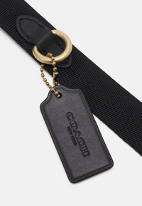 Sangle noire texturée avec un anneau en métal doré et une étiquette en cuir noire embossée "COACH New York", attachée par une petite chaîne à billes dorée.
