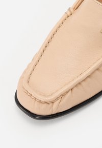 Beige lædersko med rund tå, der har synlige søm og en blød tekstur; sålen er sort og let forhøjet.