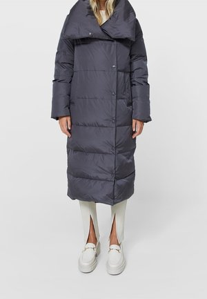 Femme portant un long manteau matelassé gris foncé avec un grand col, un pantalon blanc à ourlet fendu et des mocassins blancs, debout sur un fond uni.