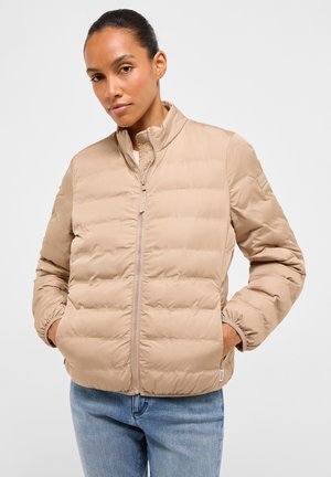 Frau trägt eine beige gesteppte Jacke mit den Händen in den Taschen und blaue Jeans, steht vor einem schlichten hellgrauen Hintergrund.