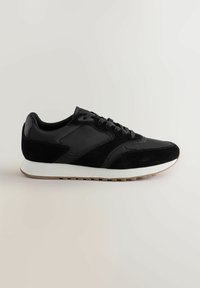 Schwarzer Sneaker mit Wildleder- und Stoffelementen, schwarzen Schnürsenkeln, weißer Zwischensohle und einer Laufsohle aus Gummigummi vor einem neutralen Hintergrund.