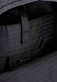 Interno nero di una borsa con una texture liscia, caratterizzato da un motivo ripetuto "Armani Exchange" e un piccolo etichetta con il logo "A/X".