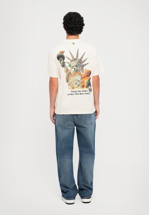 T-shirt beige chiaro con una stampa grafica colorata che raffigura una statua di gatto stilizzata e il testo: "Governando le strade di New York come un orso."