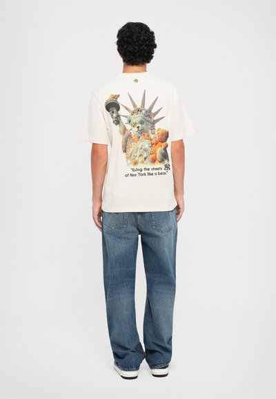 T-shirt beige chiaro con una stampa grafica colorata che raffigura una statua di gatto stilizzata e il testo: "Governando le strade di New York come un orso."