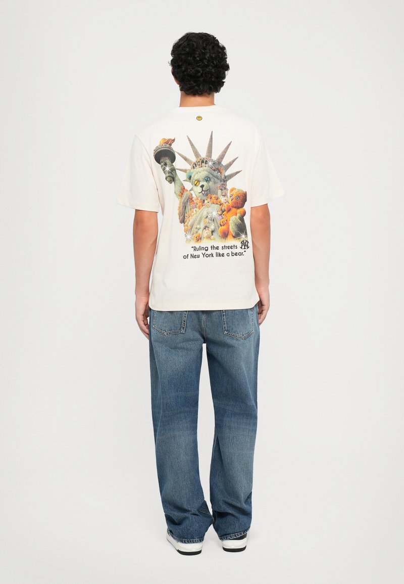 T-shirt beige chiaro con una stampa grafica colorata che raffigura una statua di gatto stilizzata e il testo: "Governando le strade di New York come un orso."