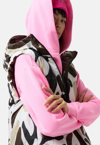 Kamizelka ocieplana w brązowo-biały wzór moro, nałożona na różową bluzę z kapturem z wydłużonymi mankietami. Model stoi z założonymi skrzyżowanymi ramionami.