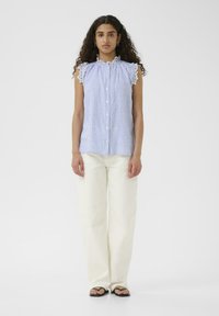 Top senza maniche a righe blu e bianche con colletto a frange e chiusura con bottoni, abbinato a pantaloni larghi beige chiaro e sandali neri.