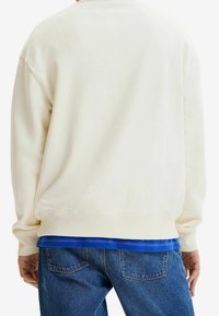Sudadera de color crema con mangas largas y puños acanalados, superpuesta sobre una camisa azul. Combinada con jeans de mezclilla azul.