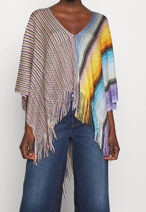 Poncho tricoté multicolore avec sections rayées et à motifs, bords frangés, porté sur un jean bleu par une personne à la peau foncée.