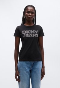 Svart bomulls-t-skjorte med "DKNY JEANS" i sølvnitter. Kort erm, rund hals, kombinert med lyseblå jeans med slitte detaljer.