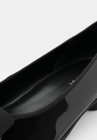 Ballerina nera in vernice con punta arrotondata, fascia elastica testurizzata sulla parte superiore e interno liscio.