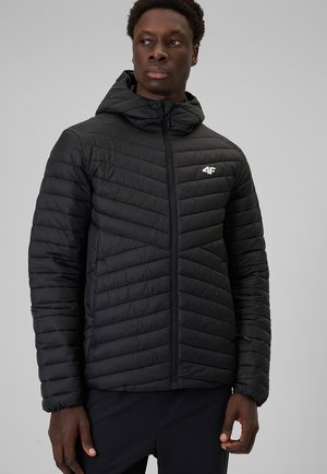 Mężczyzna ubrany w czarną pikowaną kurtkę z kapturem i zamkiem błyskawicznym, stoi na jednolitym szarym tle i patrzy na bok.