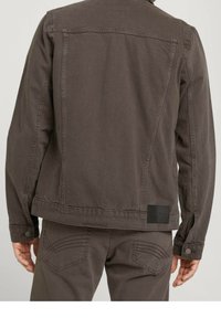 Veste en coton marron avec une coupe droite, des coutures visibles et un design arrière carré. Elle présente une étiquette noire sur le côté inférieur droit.
