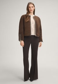 Brązowa bomberka z polaru z detalami na suwak, noszona na kremowym, dzianym swetrze. Czarna rozkloszowana spodnie i czarne, spiczaste buty uzupełniają strój.