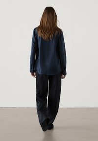 Camicia blu navy con maniche lunghe, tessuto morbido, colletto a punta, vestibilità ampia, abbinata a pantaloni scuri larghi, vista da dietro.
