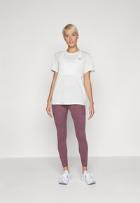 Balta trumpų rankovių marškiniai su subtiliu mėlynu logotipu, derinami su prigludusiomis mauve spalvos sportinėmis kelnėmis ir baltomis sportinėmis bateliais, prieš paprastą foną.