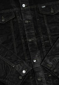 Schwarze Cordjacke mit silbernen Knöpfen, gestickten Details und zwei Brusttaschen. Strukturierter Stoff und dezente gelbe Akzente.