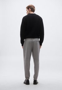 Fekete kötött pulóver, szürke szabadidőnadrág rugalmas mandzsettákkal, és fekete cipők. Hátrafelé nézve, egyszerű világos háttér előtt.