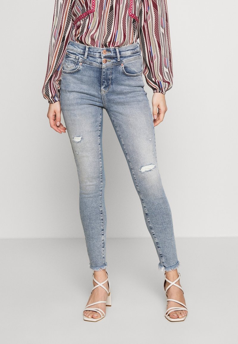 Mujer que lleva jeans ajustados desgastados de color azul claro con dobladillo sin terminar, blusa de manga larga con estampado y sandalias de tacón cuadrado blanco con tiras.