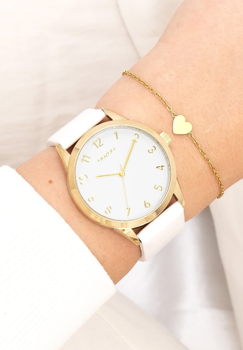 Montre circulaire dorée avec bracelet en cuir blanc, dotée d'un cadran blanc et d'index d'heures en or. Accentuée par une fine chaîne dorée avec un charm en forme de cœur.