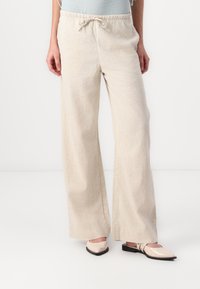 Pantaloni a gamba larga realizzati in tessuto beige testurizzato, con vita elastica e cordoncino. Indossati con scarpe basse di colore rosa chiaro.