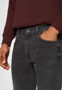 Ein Mann trägt dunkelgraue Lee-Jeans, mit einer Hand in der vorderen Tasche, und ein weinrotes Langarmshirt, das an der Taille und an den Ärmeln sichtbar ist.