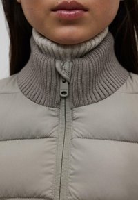 Gilet imbottito grigio con collo alto a costine. Presenta una zip frontale e una texture liscia e trapuntata. Il materiale sembra caldo e resistente.