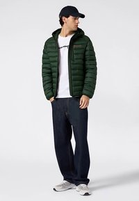 Groene pufferjas met capuchon, geribbelde textuur, ritssluiting aan de voorkant, gedragen over een wit T-shirt. Gecombineerd met donkerblauwe spijkerbroek en grijze sneakers.