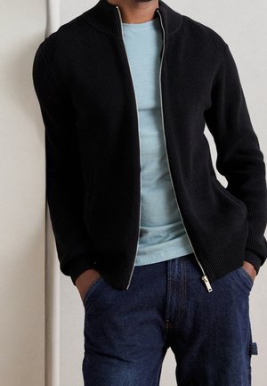 Mężczyzna ubrany w czarny sweter na zamek z dzianiny na jasnoniebieską koszulę, z rękami w kieszeniach niebieskich dżinsów.