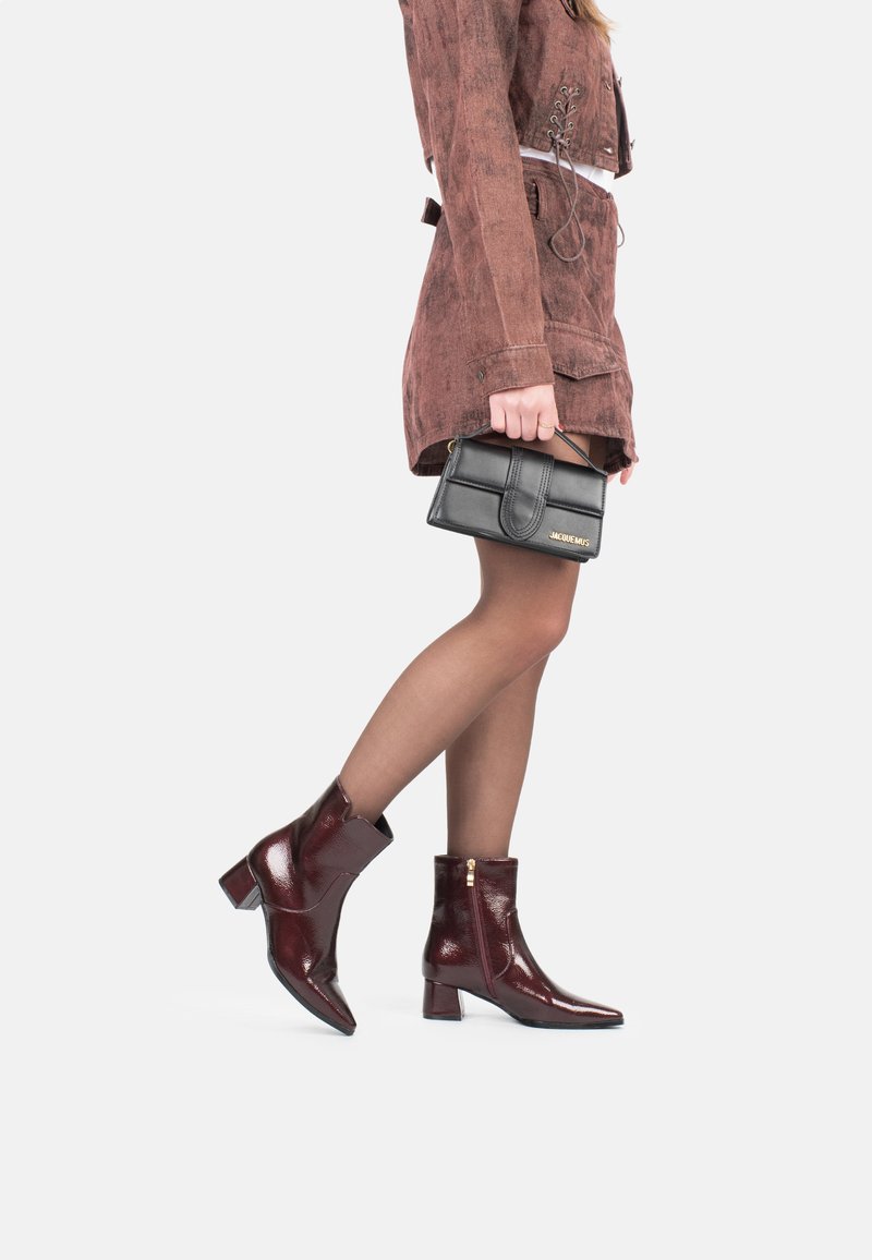 Botines de cuero burdeos con un acabado brillante, tacones de bloque y cremalleras en los laterales. Combinados con un vestido marrón texturizado y un bolso negro.