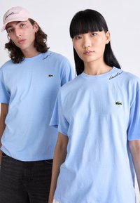 Két fiatal felnőtt világoskék Lacoste pólót visel, az egyikük pink Lacoste sapkát hord. Egymás mellett állnak egy fehér háttér előtt.