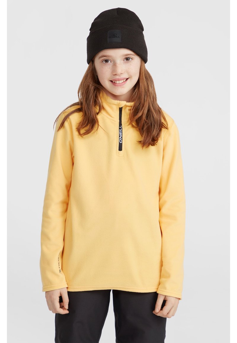 Gelber Fleece-Pullover mit einem Viertel-Reißverschluss, schwarzen Reißverschlussdetails und langen Ärmeln. Getragen mit einer schwarzen Strickmütze. Glatte Textur.